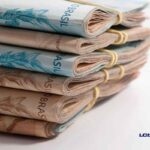 Maços de notas de reais dobradas e presas com elásticos, empilhadas sobre uma superfície branca, com o logo do LCbank no canto inferior direito