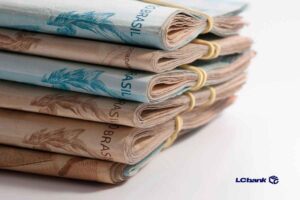 Maços de notas de reais dobradas e presas com elásticos, empilhadas sobre uma superfície branca, com o logo do LCbank no canto inferior direito